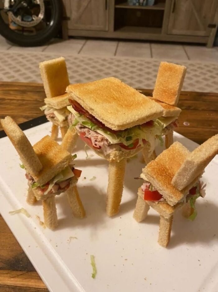 Miniature table and chairs made entirely from stacked sandwiches with toasted bread and deli fillings on a white tray.