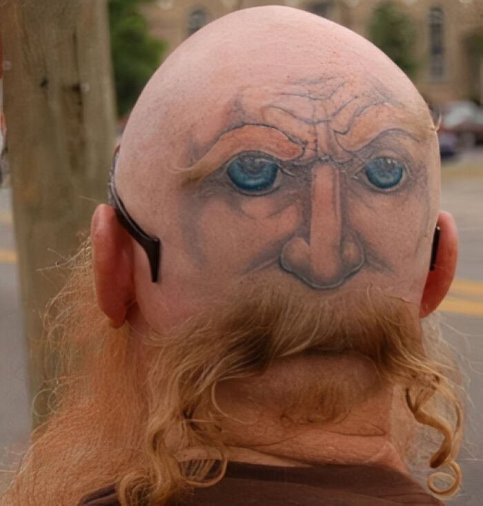 Tattoo of a face with blue eyes and furrowed brows inked on the back of a bald head with a red beard below.