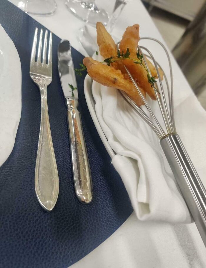 Fried food served on a metal whisk with herbs, next to fork and knife on a napkin, showing unholy meal presentation.
