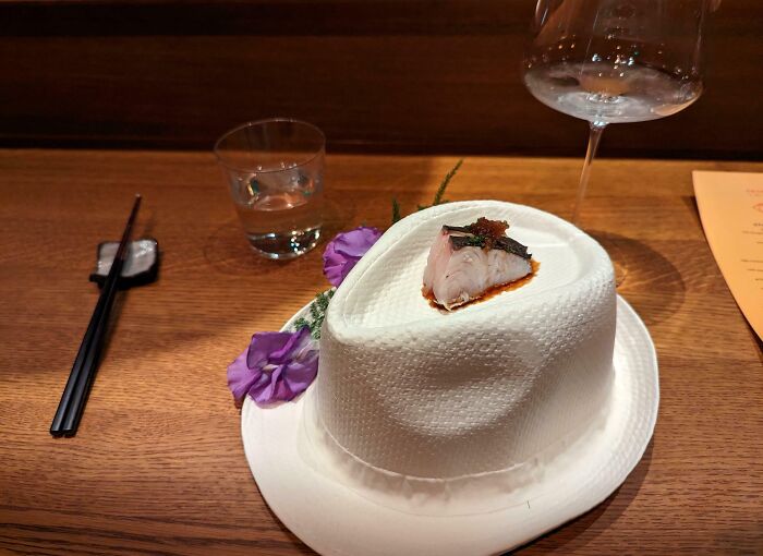Unholy meal presentation with fish served on a textured white hat-shaped plate at a wooden table setting.