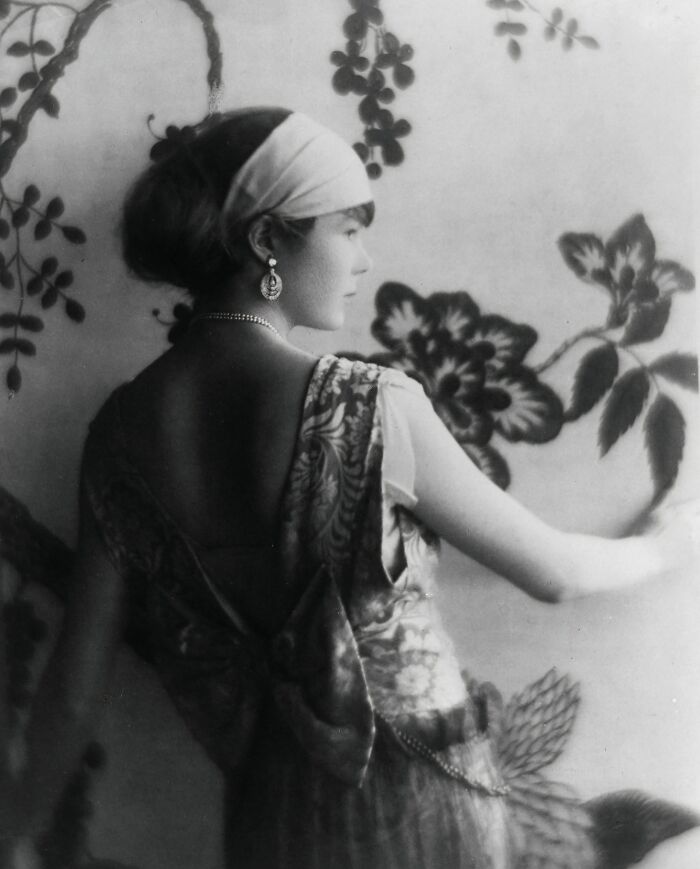 Black and white photo of a woman from the 1900s wearing a headband and ornate dress standing by a floral patterned wall.