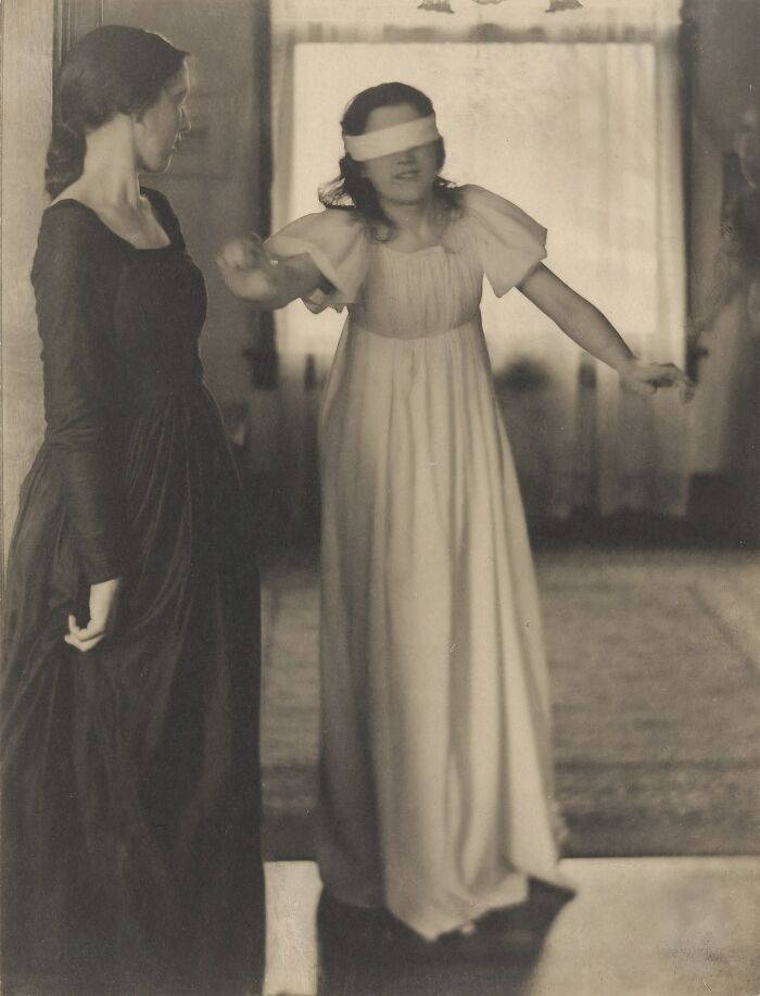 Two women from the 1900s, one blindfolded and reaching out while the other watches in vintage attire showing beauty of women.