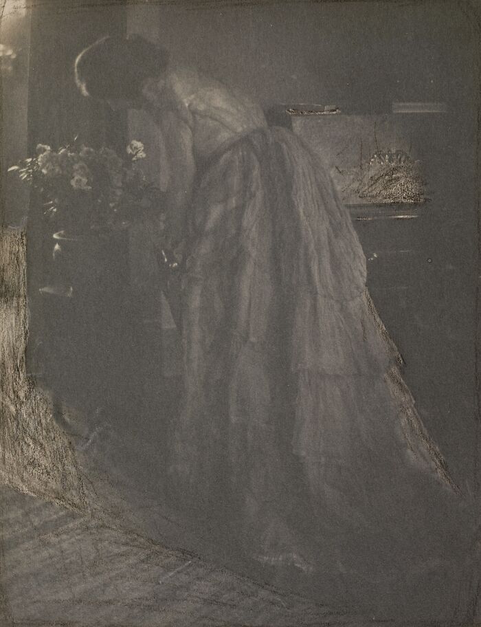 Vintage photo of a woman in a long dress bending near a flower vase, showcasing the beauty of women from the 1900s.