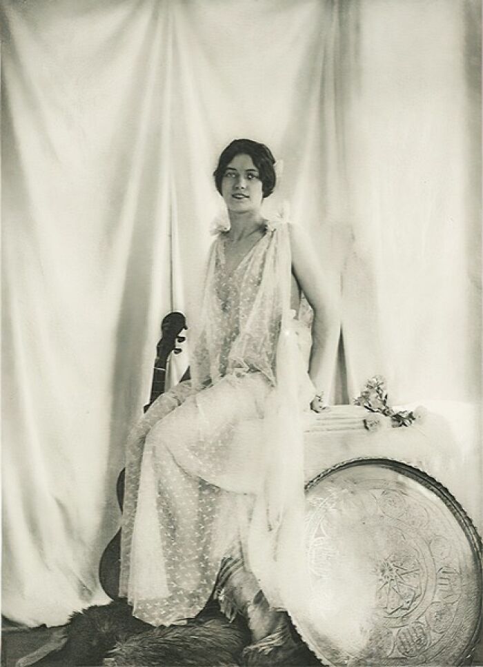 Vintage photo of a woman from the 1900s wearing a sheer dress, seated beside a guitar and an ornate shield.