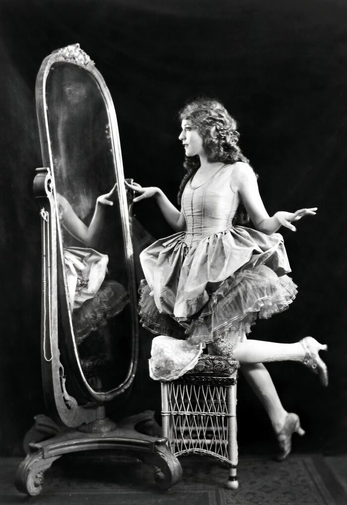 Vintage photo of a woman from the 1900s in a tutu dress posing gracefully by an ornate standing mirror.