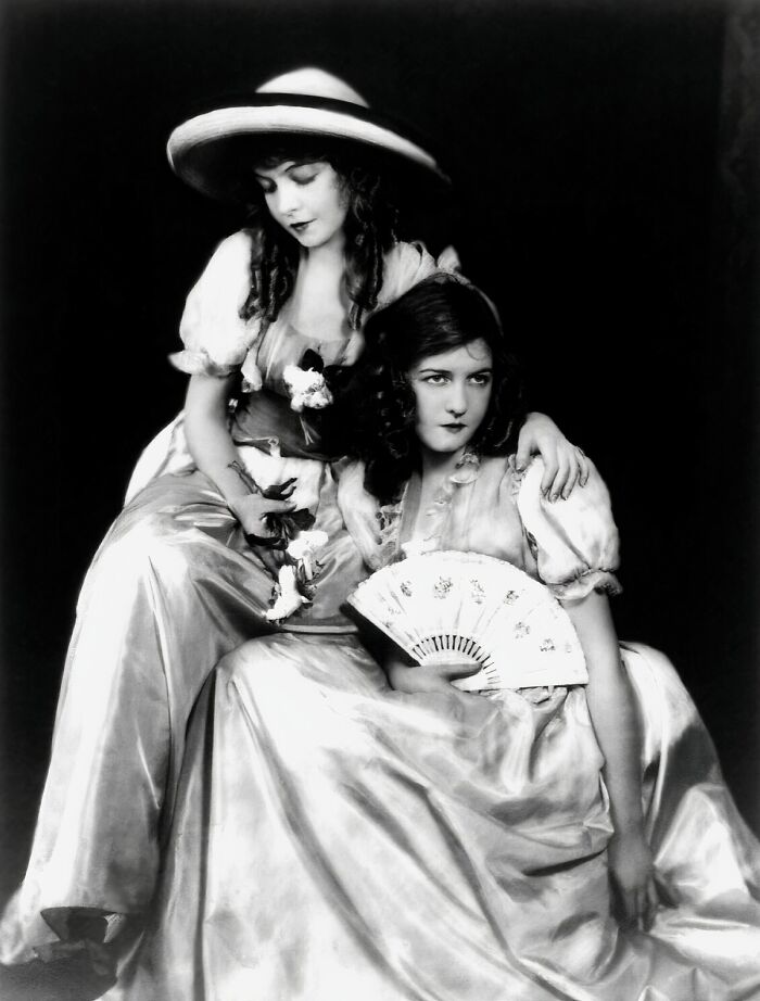 Two women from the 1900s in elegant vintage dresses, one holding a fan, showcasing classic beauty and style.
