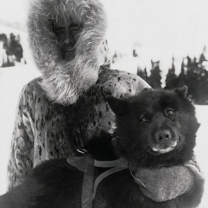 Man in fur parka holding a large dark sled dog in snowy wilderness, unique old photo capturing historical moment.