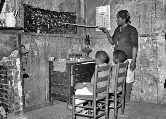 Unique old photo of a woman teaching children in a rustic classroom with handwritten lessons on a chalkboard.
