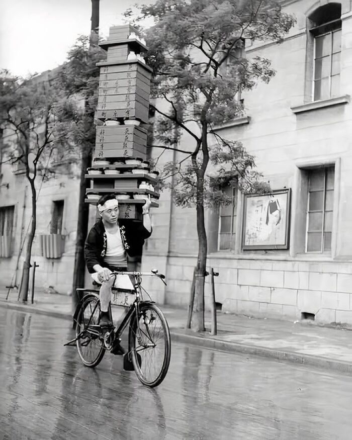 Man balancing tall stack of boxes on his head while riding a bicycle on a wet street in a unique old photo.