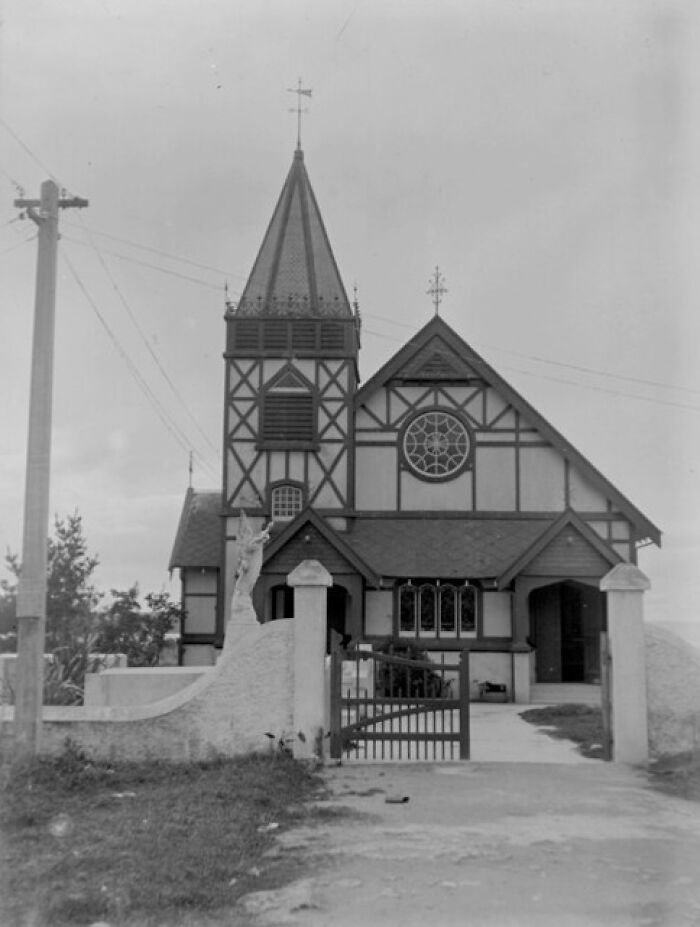 Black and white historical photograph of a church with a tall steeple and gated entrance from 100 years ago.