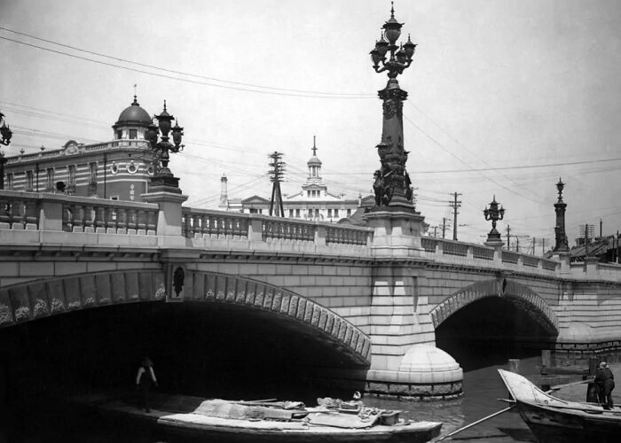 Black and white historical photograph of a stone bridge with ornate lampposts over a river, showing the world 100 years ago.