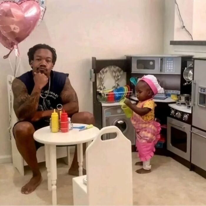 Man with tattoos sitting thoughtfully at a small table as a child plays with a toy kitchen in a wholesome, amazing setting.