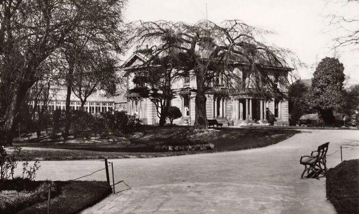 Black and white historical photograph of a large mansion surrounded by trees and benches, showing the world 100 years ago.