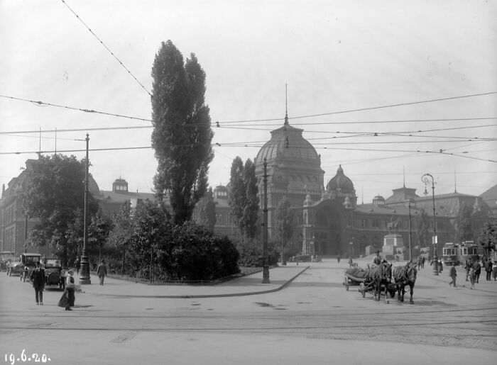 Black and white historical photograph of a city street with horse-drawn carriages and early automobiles from 100 years ago.