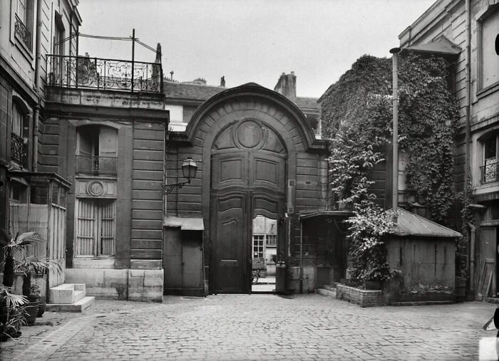 Historic black and white photograph showing a large arched wooden gate in a cobblestone courtyard from 100 years ago.