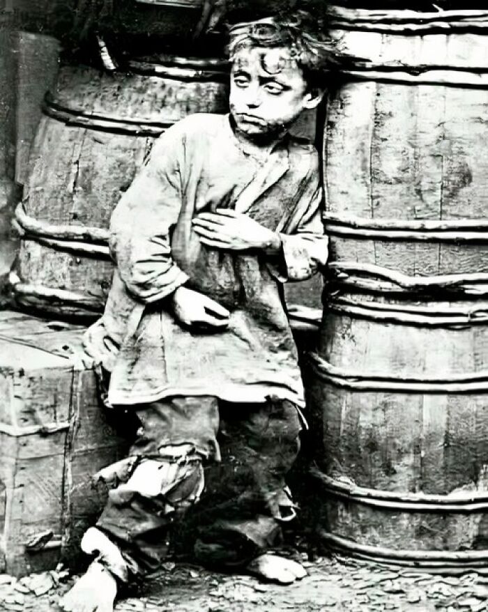 Black and white old photo of a barefoot child with torn clothes leaning against wooden barrels showing unique historical life.