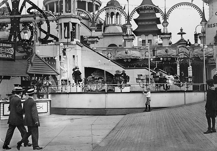 Vintage amusement park scene with people walking and old carousel ride in rare and interesting historical photograph.
