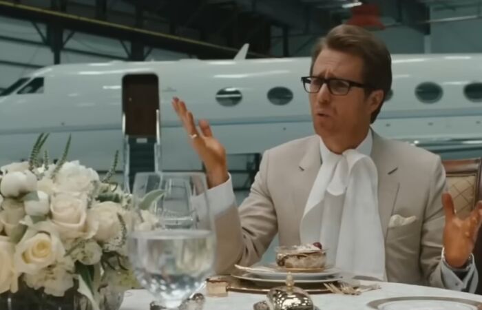 Actor in a beige suit and glasses gesturing at a dining table set inside a hangar with a private jet behind in iconic villains scene.