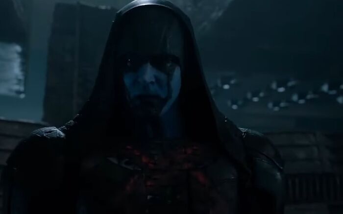 A dark, hooded villain with a painted blue face appearing in a dimly lit, ominous setting, iconic villain portrayal.