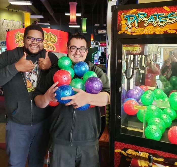 Two men smiling by a claw machine, one holding colorful balls, showing amazing luck at an arcade game.