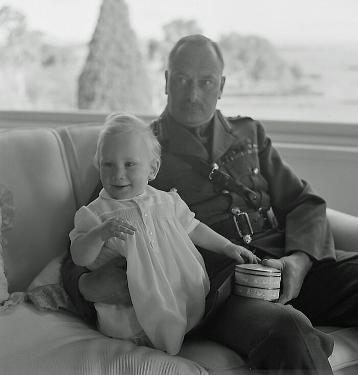 Black and white rare historical photograph of a man in military uniform holding a smiling toddler on a couch indoors.