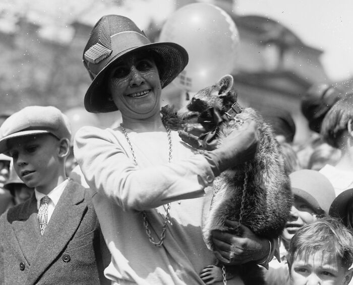 Woman smiling while holding a raccoon, surrounded by children in a rare and interesting historical photograph.