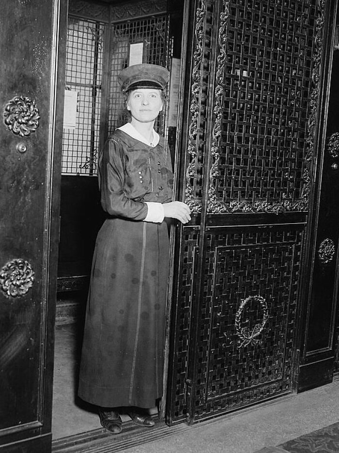 Woman in early 20th century attire operating an ornate elevator door in a rare and interesting historical photograph.