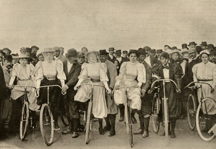 Women in vintage dresses preparing to ride bicycles with a crowd watching in a rare historical photograph.