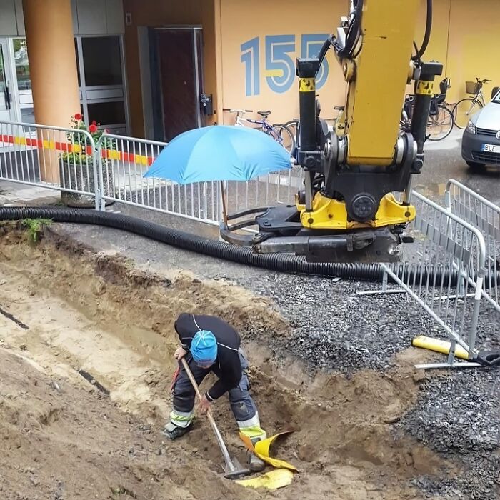 Worker digging in a trench under a blue umbrella held by construction machinery, showing wholesome people and animals care.