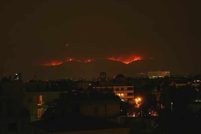 Nighttime view of catastrophic wildfires burning across hills near a city, with glowing flames and smoke in the sky.