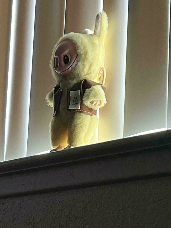 Creepy Labubus fake doll with a distorted face standing on a windowsill with backlighting from vertical blinds.