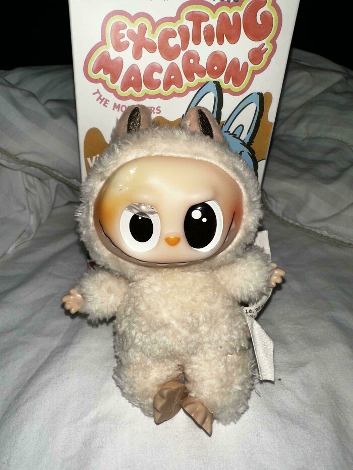 Creepy Labubus doll with large eyes and animal costume, one eye damaged, sitting on bed with Exciting Macaron box behind.