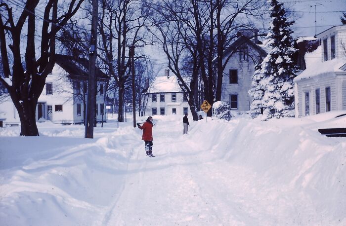 Children playing in a snowy street between houses, capturing daily life across the globe in the 50s winter season.