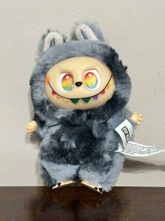 Plush creepy Labubus figure with rainbow eyes and sharp colorful teeth, standing on a wooden surface.