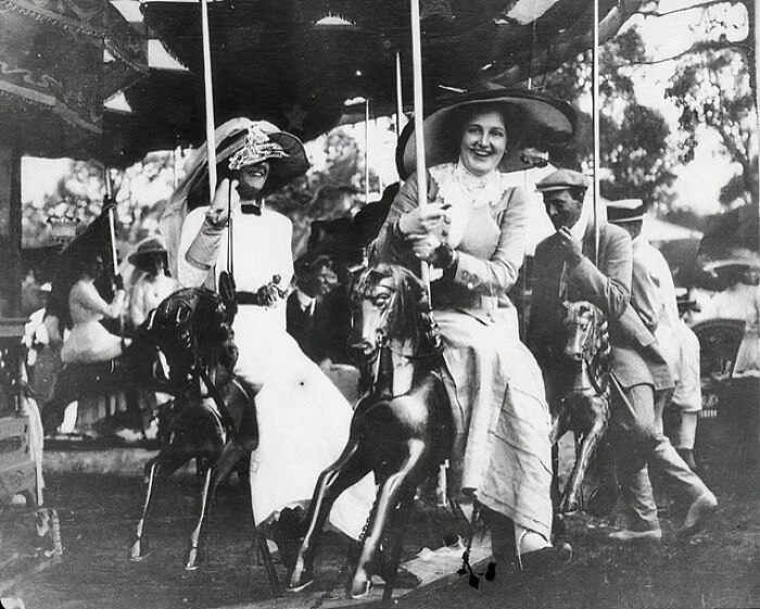 Two women in early 1900s attire smiling and riding a vintage carousel in a rare historical photograph.