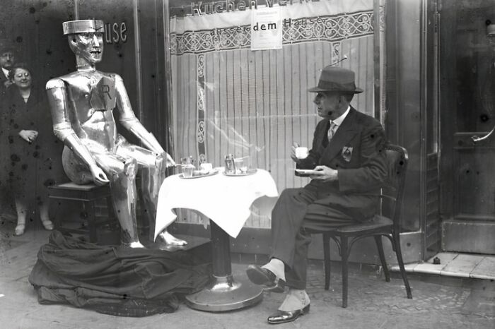 Man in suit and hat enjoys coffee at table with a large silver robot, a rare and interesting historical photograph.