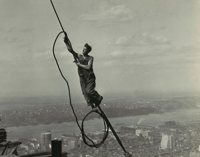 Man balancing on high steel beam with cables above cityscape in rare and interesting historical photograph.