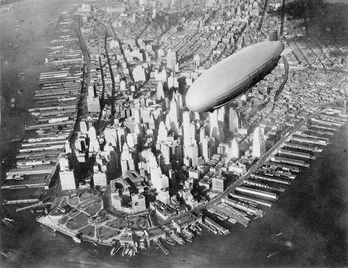 A rare historical photograph showing a large zeppelin flying over a densely packed city skyline by the waterfront.