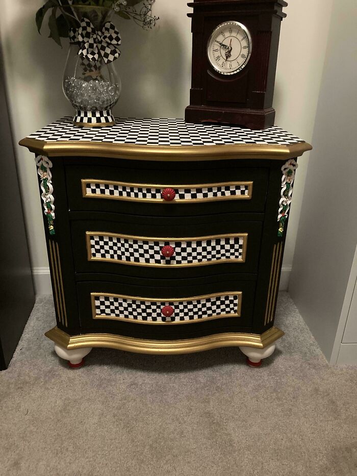 Black and gold checkerboard furniture flipping project with red knobs and floral accents on a three-drawer cabinet in a room corner.