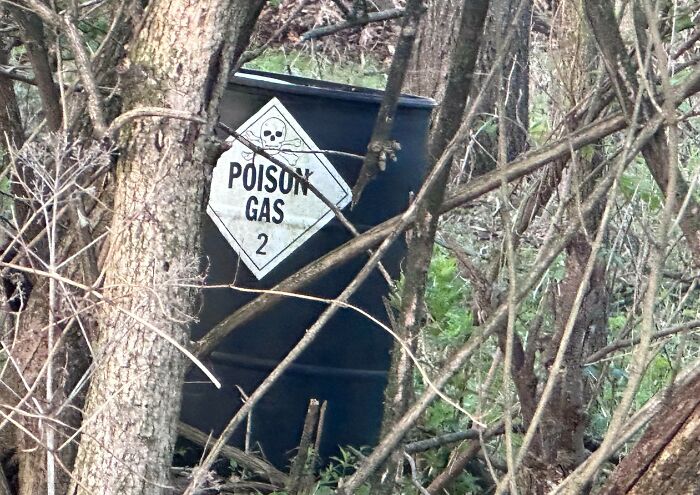 Black barrel with poison gas warning symbol hidden among trees and branches in a forest setting.