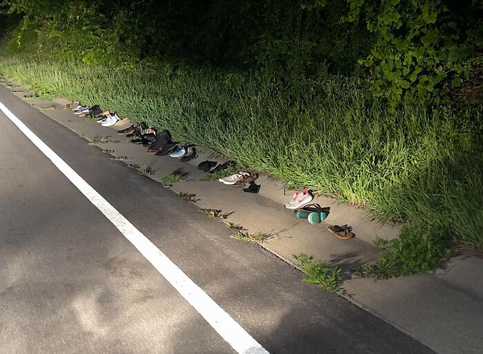 Multiple pairs of abandoned shoes lined up by a road near forest grass under dim lighting, suggesting creepy forest findings.