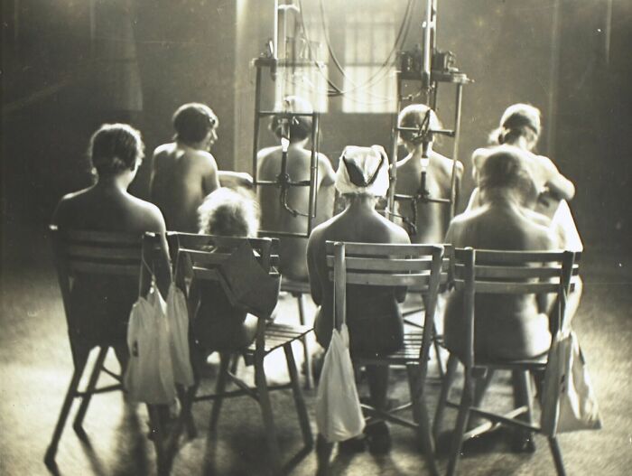 Group of patients seated in chairs with unsettling medical devices attached to their backs in a vintage clinical setting.