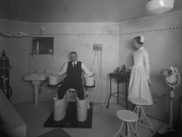 Man undergoing treatment with unsettling medical devices from the past while a nurse observes in a vintage clinical room.