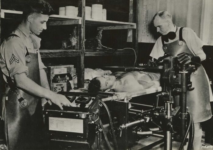 Two men operate unsettling medical devices on a patient lying on a table in a vintage medical setting.