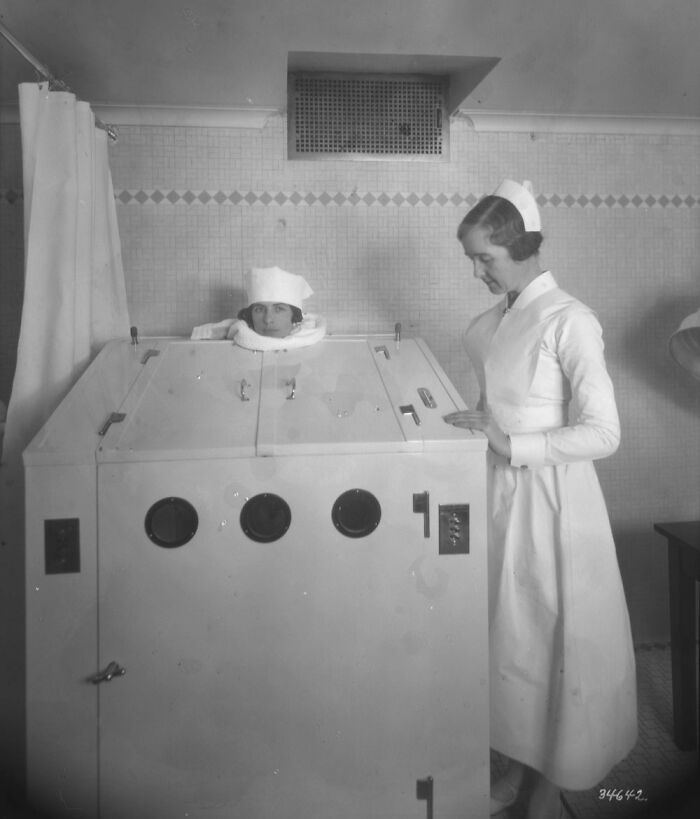 Vintage medical device with patient inside a large enclosed chamber, supervised by a nurse in early 20th century attire.