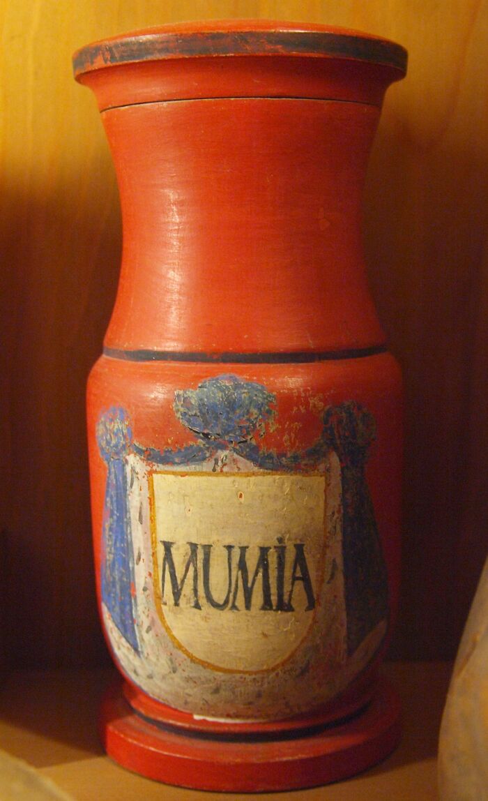 Old red medical container labeled MUMIA, an unsettling medical device from the past displayed on a wooden shelf.