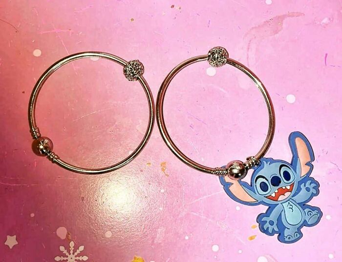 Two silver bracelets on a pink surface, one featuring a blue cartoon character charm resembling a glitch in the matrix scene.