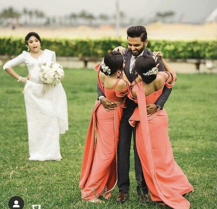 Groom hugging two bridesmaids in orange dresses outdoors while the bride in white looks shocked at the scene.
