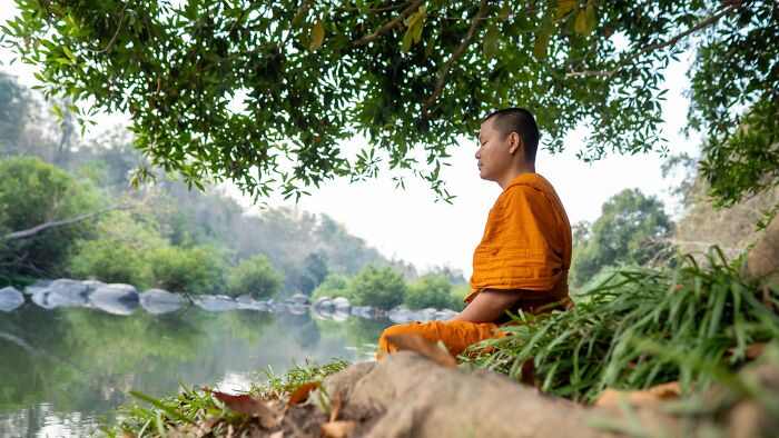 Buddhist monk meditating by a river surrounded by green trees, reflecting calmness beyond common religious myths.