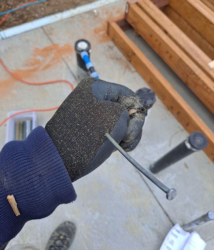 Gloved hand holding a nail perfectly balanced through a worn spot, showing unbelievable luck at a construction site.
